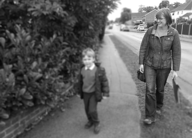 A Mum walking with her son to school. The pair look happy and content to be walking to school together. The mum is carrying her son's school book bag. She is smiling and looking proudly at her son. Her son looks relaxed and confident as he walks beside her. He is dressed smartly in his uniform, ready for school. 