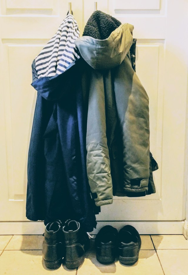A Parent's coat is hanging on the door handle next to a child's coat. Beneath the coats are the parents boots and the child's shoes. Both the parent's and the child's coats and shoes are easy to access and ready to put on before leaving the house to go to school.