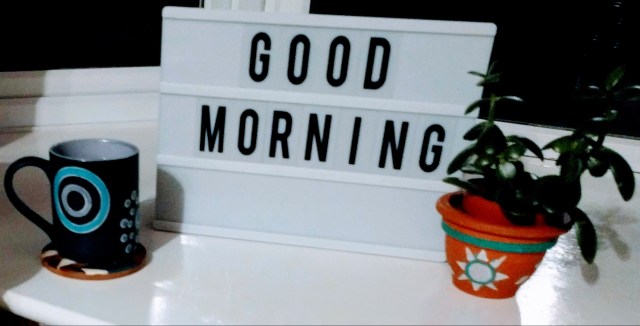 The photo shows a calm scene. A letters board has been completed with the words "good morning", all set up ready to greet the early riser who has achieved their goal of waking up earlier. The letters board is on a window ledge in between a plant placed on the right hand side and a drinks mug, placed on a coaster and waiting to be enjoyed, on the left hand side.