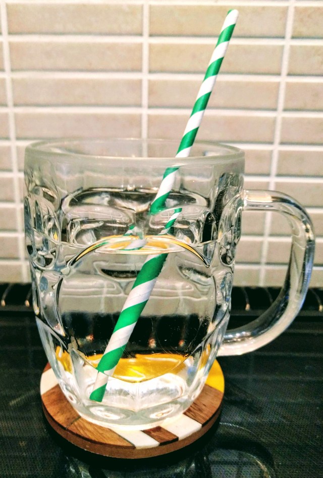 A pint glass (of a traditional design with a handle - the kind typically used for serving beer) sitting on a colourful coaster on a kitchen worktop. The pint glass is filled with water and topped with a green and white striped drinking straw. The drink of water looks visually appealing and refreshing. It shows that with a little bit of thought and intention, something as simple as a glass of water can be made to feel more like a treat to help you create a good daily habit.
