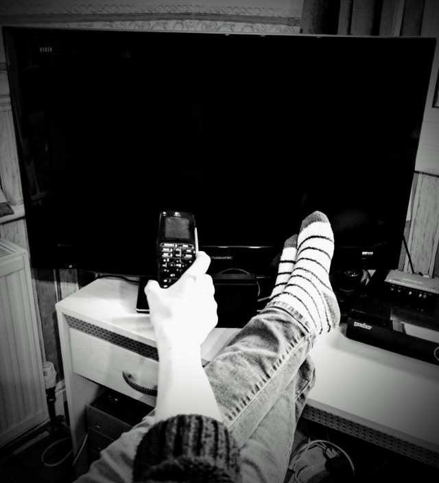 There is a large telvision screen. In front of the television screen are a persons outstretched legs resting on the television stand. The person has cozy casual socks on their feet and their feet are crossed. The person has their arm outstetched towards the T.V. screen and is holding a T.V. remote control pointing at the screen. It looks as though the person is lounging in front of the television and is either about to change the channel or switch the T.V. on/off.