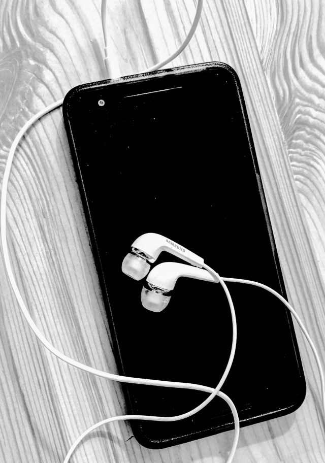 A smart phone resting on a table with some ear-buds plugged in ready for use. This shows how you can set your phone up to listen to Podcasts while you are on the go around the house or if you wish to step out into the garden.