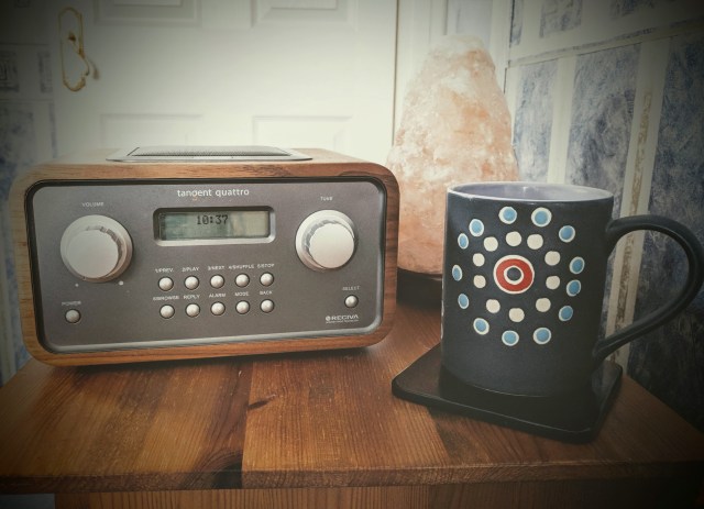 A bedside table set up with a radio alarm clock, a mug of coffee on a coaster and a Hymalayan salt lamp poistioned in the back corner. This shows a bedside table all set up for someone to drink a cup of coffee, set an alarm, listen to the radio and settle down for a short daytime nap.